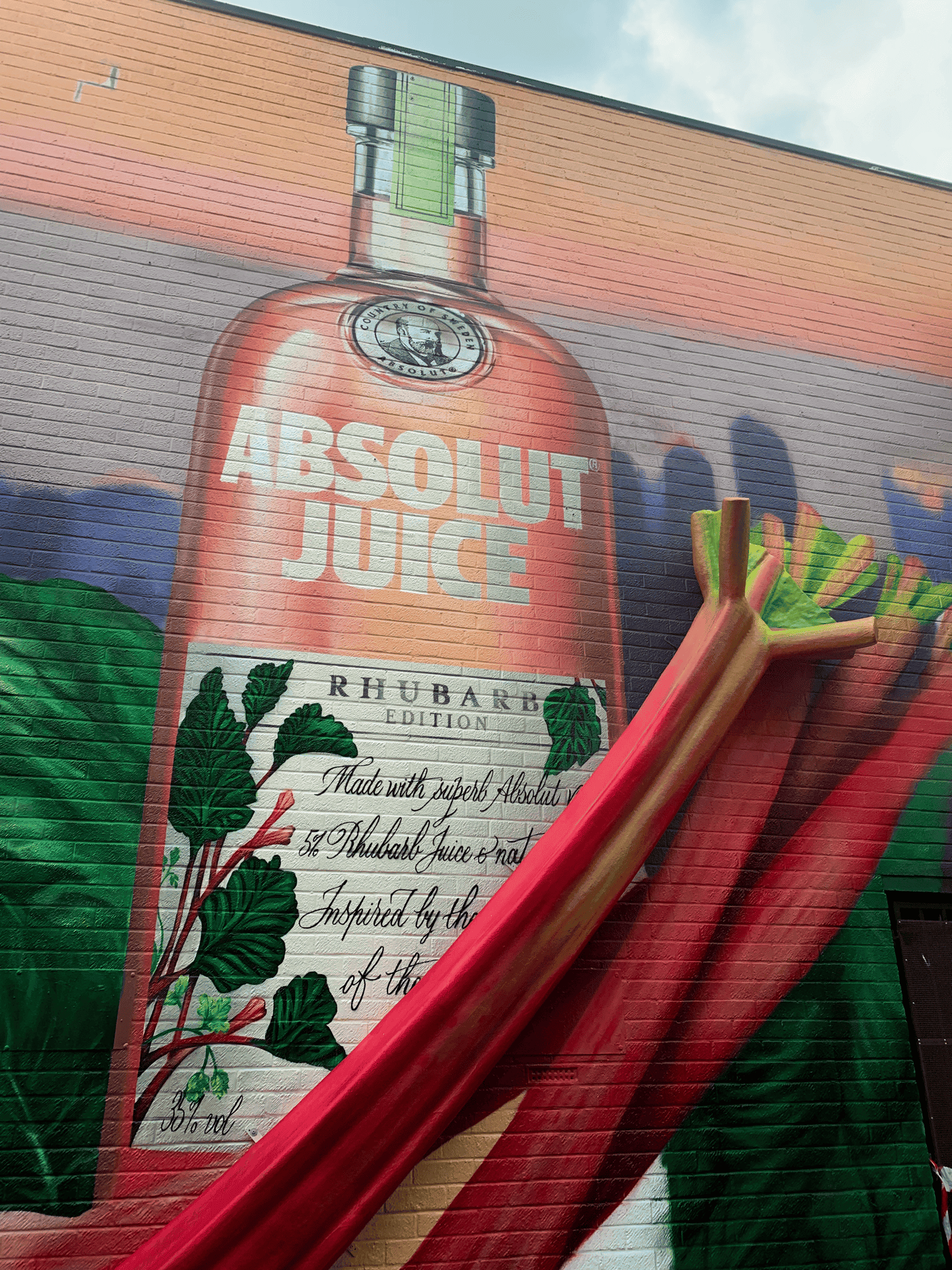 Mural of a large pink Absolut Juice Rhubarb Edition bottle on a brick wall, with vivid rhubarb stalks. Lush greenery adds a fresh, vibrant tone.