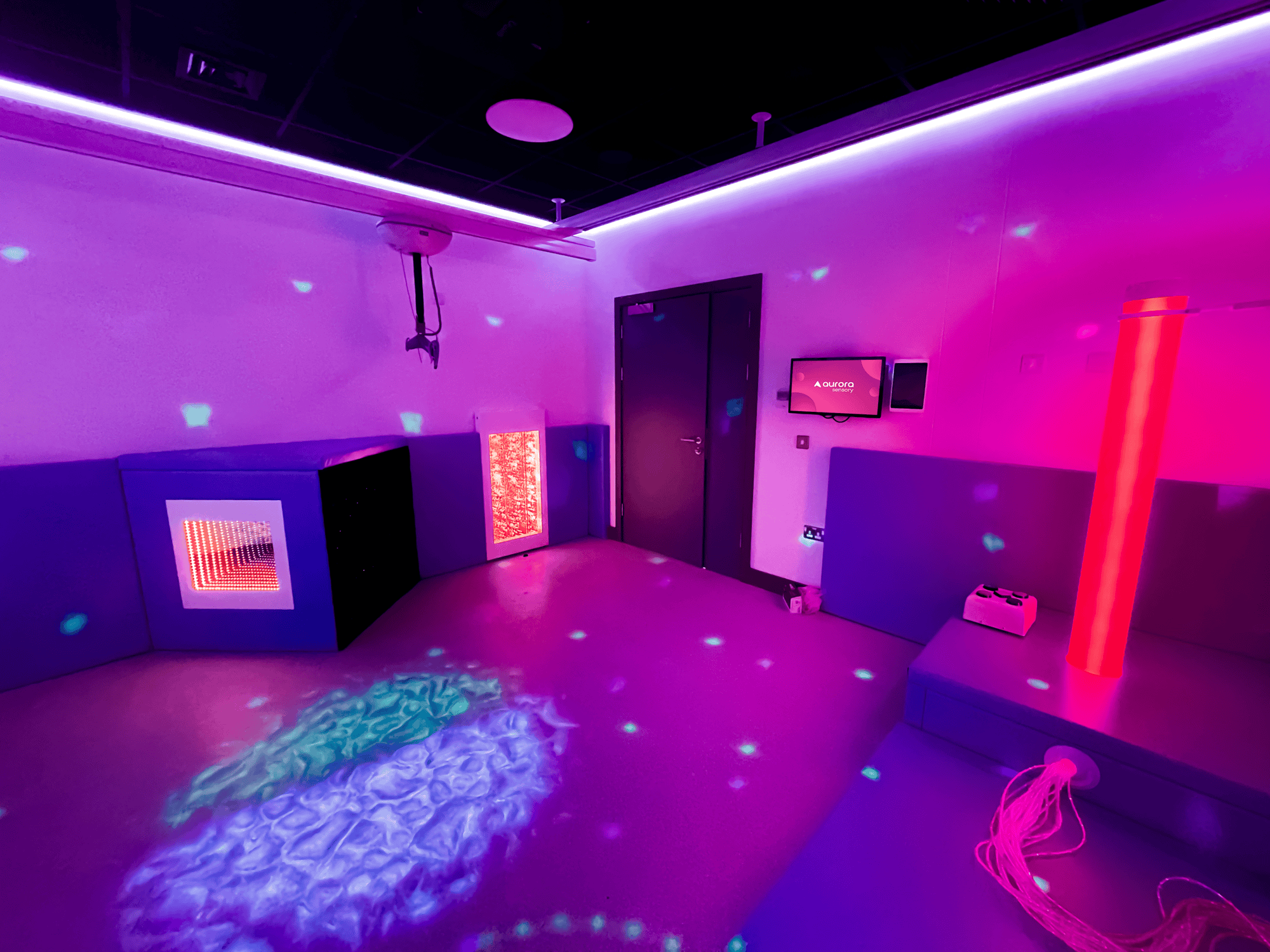 A sensory room with dim lighting and colourful projections on walls and floor. Features include a glowing bubble tube, interactive panels, and soft seating, creating a calming atmosphere.