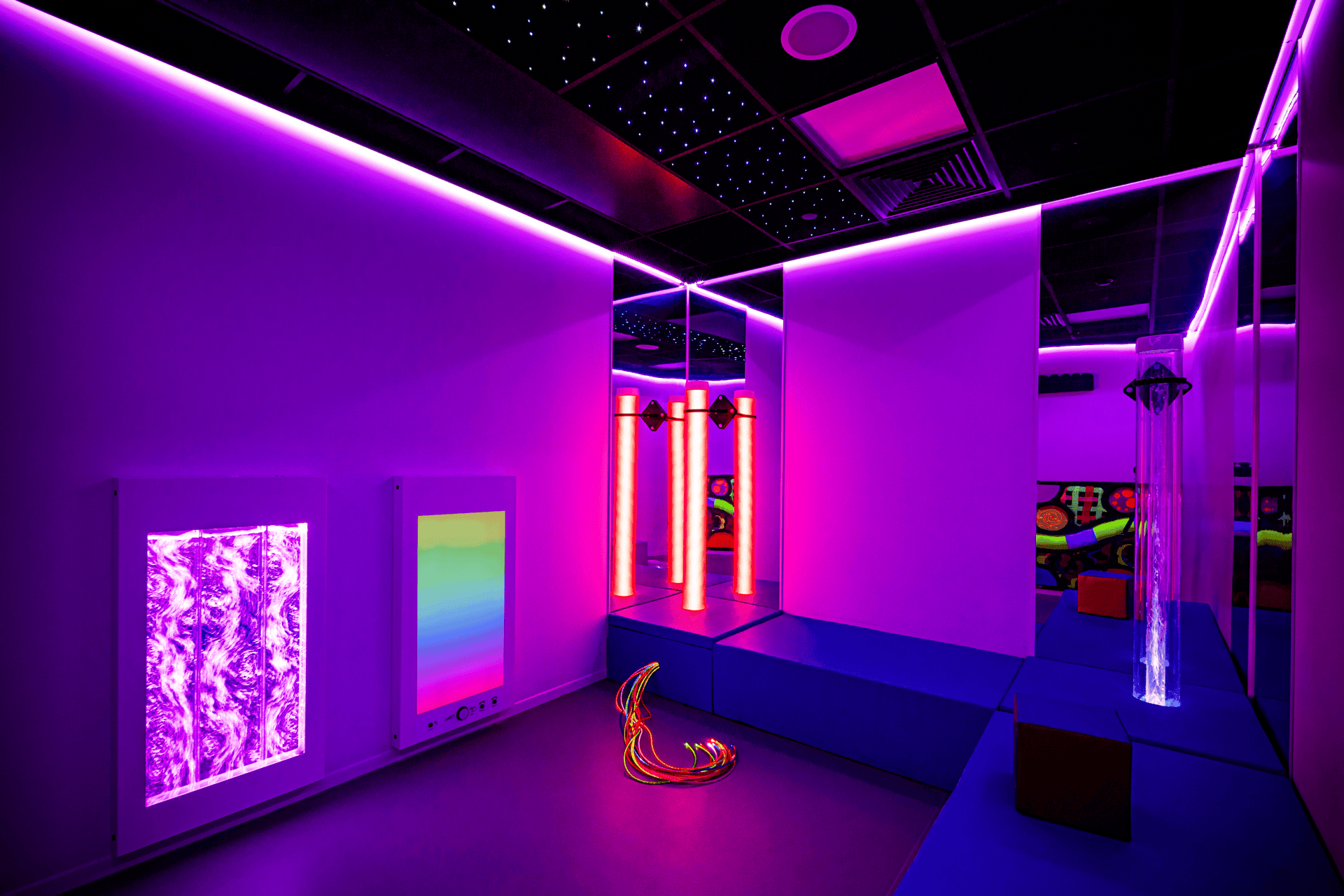 A sensory room with vibrant purple lighting, featuring luminescent bubble tubes, soft seating, mirrored walls, and colorful light panels, creating a calming atmosphere.