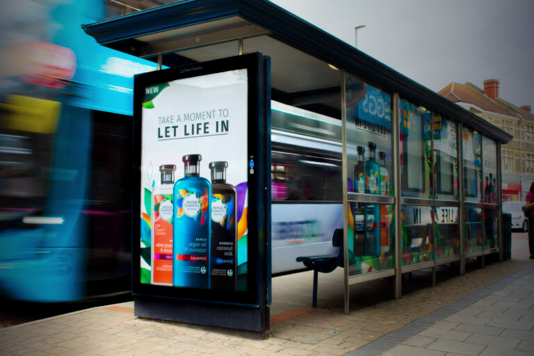 Bus stop with a billboard advertising colourful shampoo bottles and aurora hidden scent diffuser system. A blurred bus passes by, creating a dynamic, energetic urban scene.