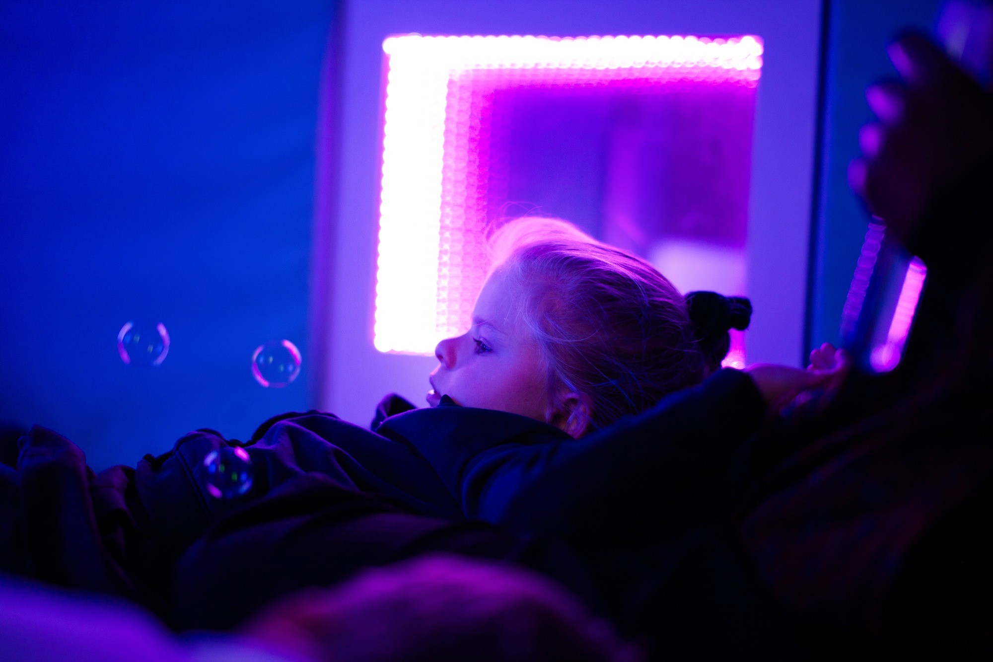 A child with blonde hair gazes at floating bubbles in a dimly lit sensory room with a soft, violet glow, creating a calming and dreamy atmosphere.