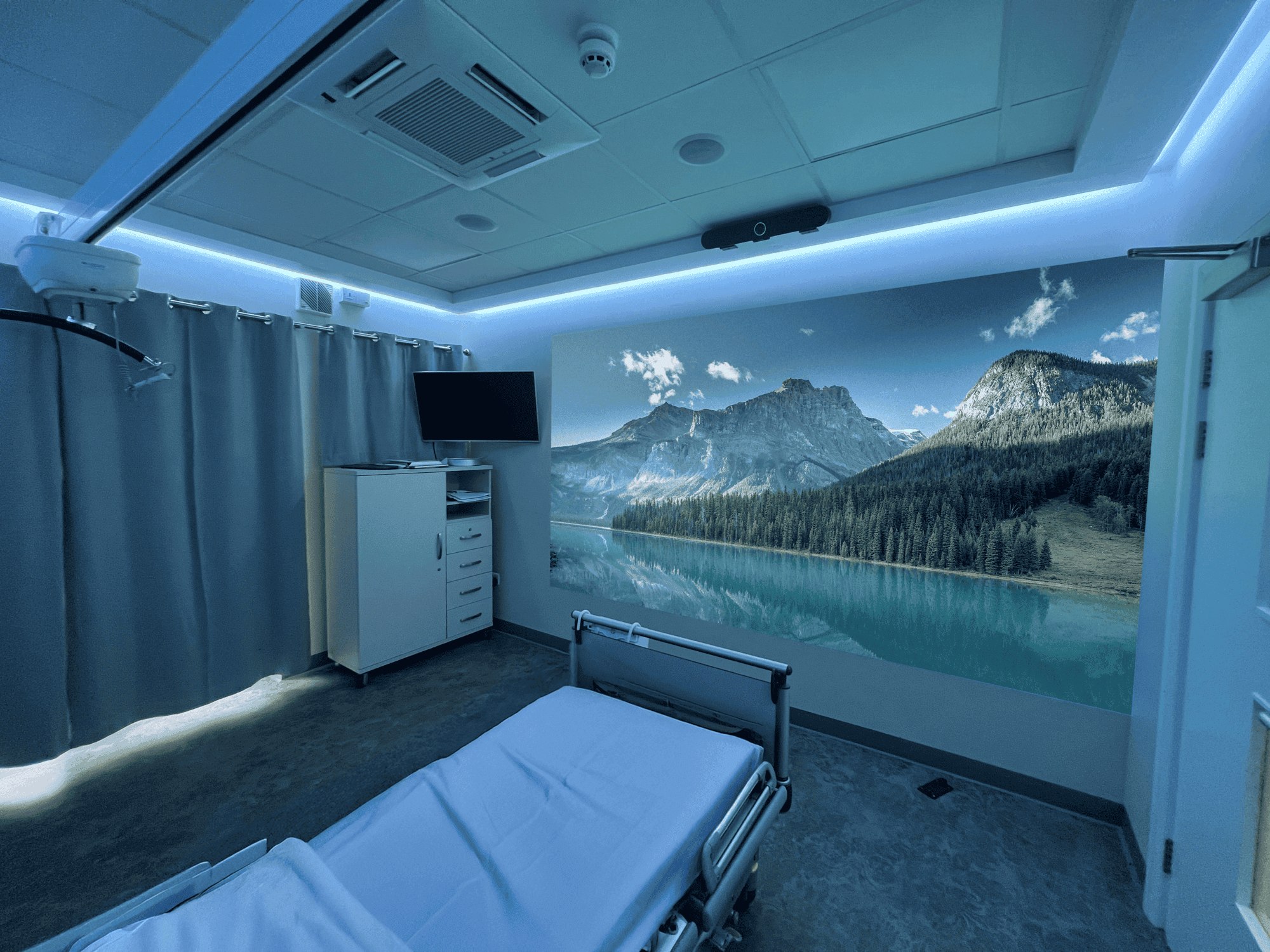 Hospital room with a white bed, grey curtains, hidden ambient coloured lighting and a single wall aurora projection image of a nature mural of mountains and a lake creates a calming atmosphere.