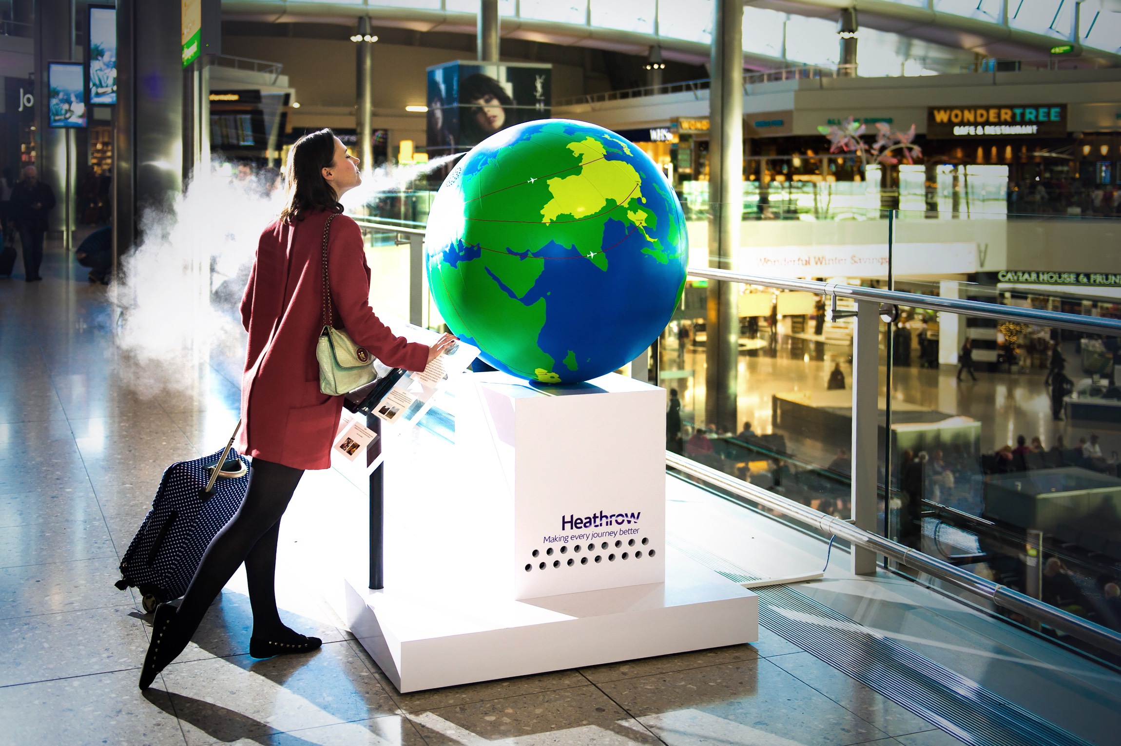 A woman in a maroon coat gazes at a large globe diffusing scent in a busy airport terminal. Sunlight illuminates the scene, creating a lively atmosphere.