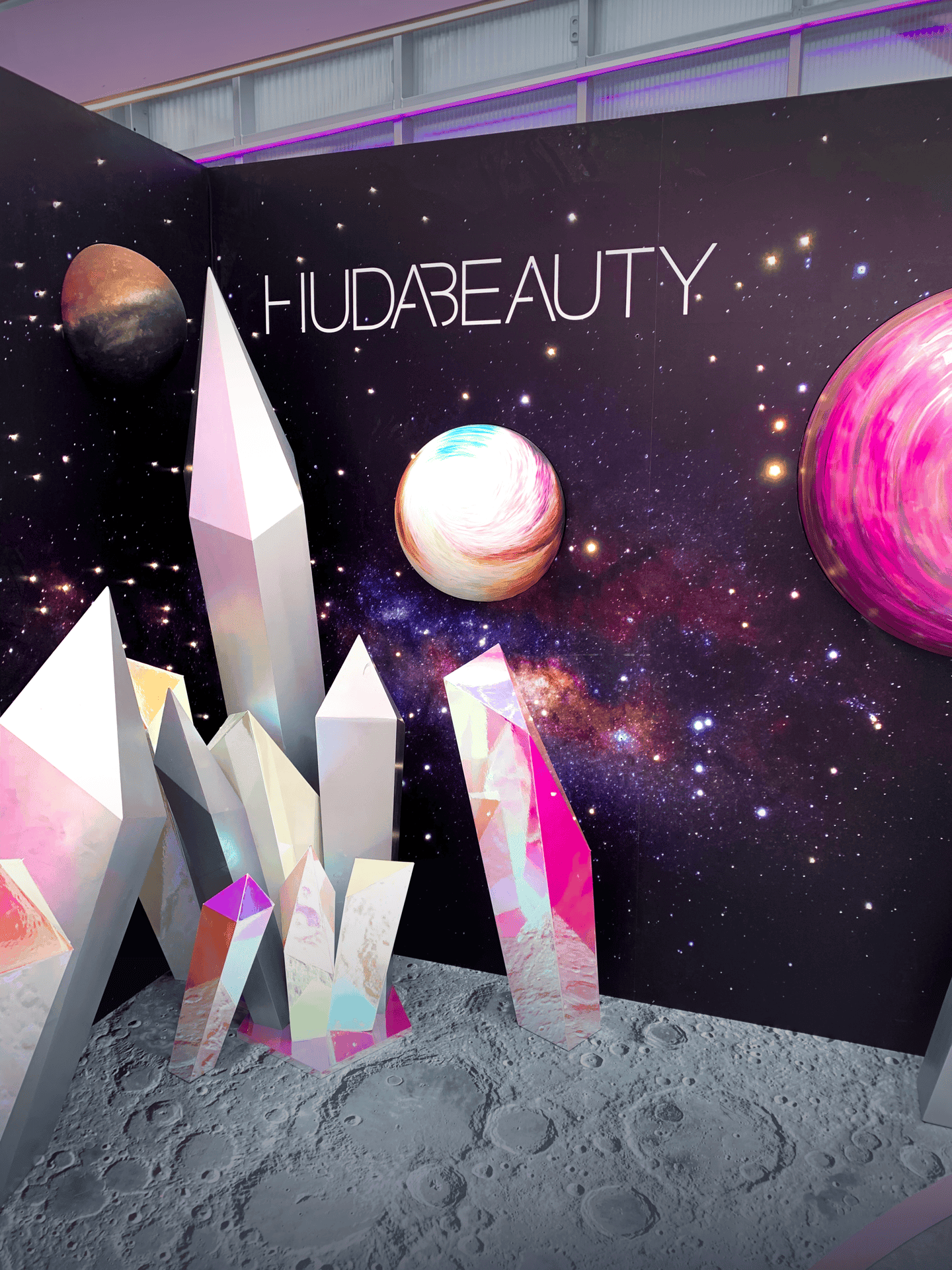 Futuristic Huda Beauty display with giant crystals on a lunar surface, cosmic background, and planets, conveying a mystical, space-themed atmosphere.