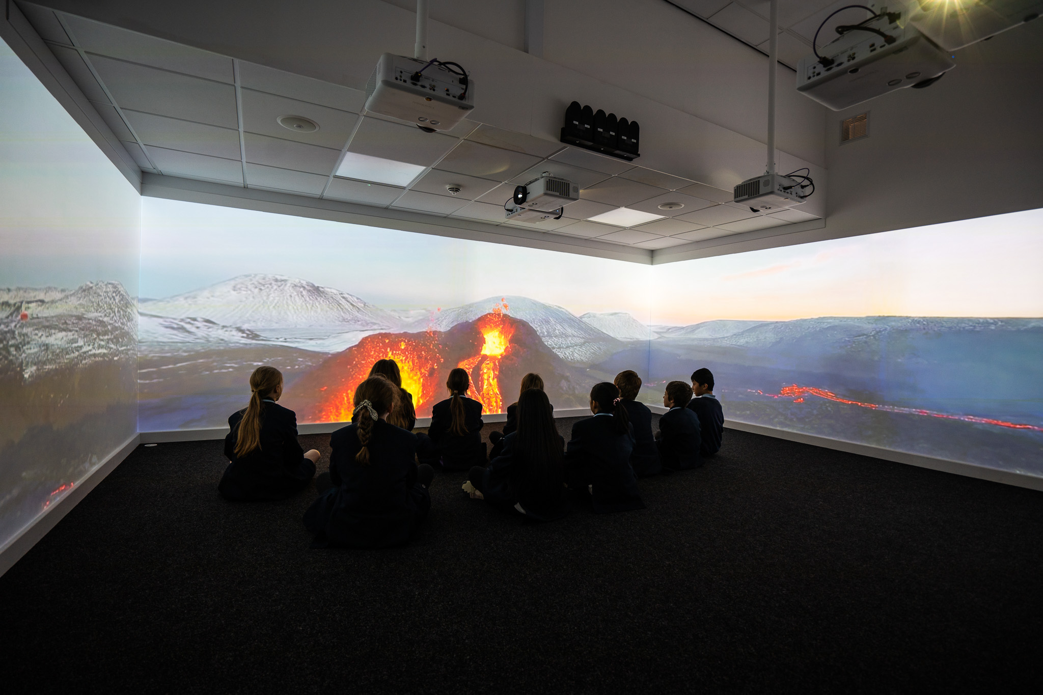Children in school uniforms sit on the floor, captivated by a vibrant projection of an erupting volcano in a darkened room, creating an immersive learning atmosphere.