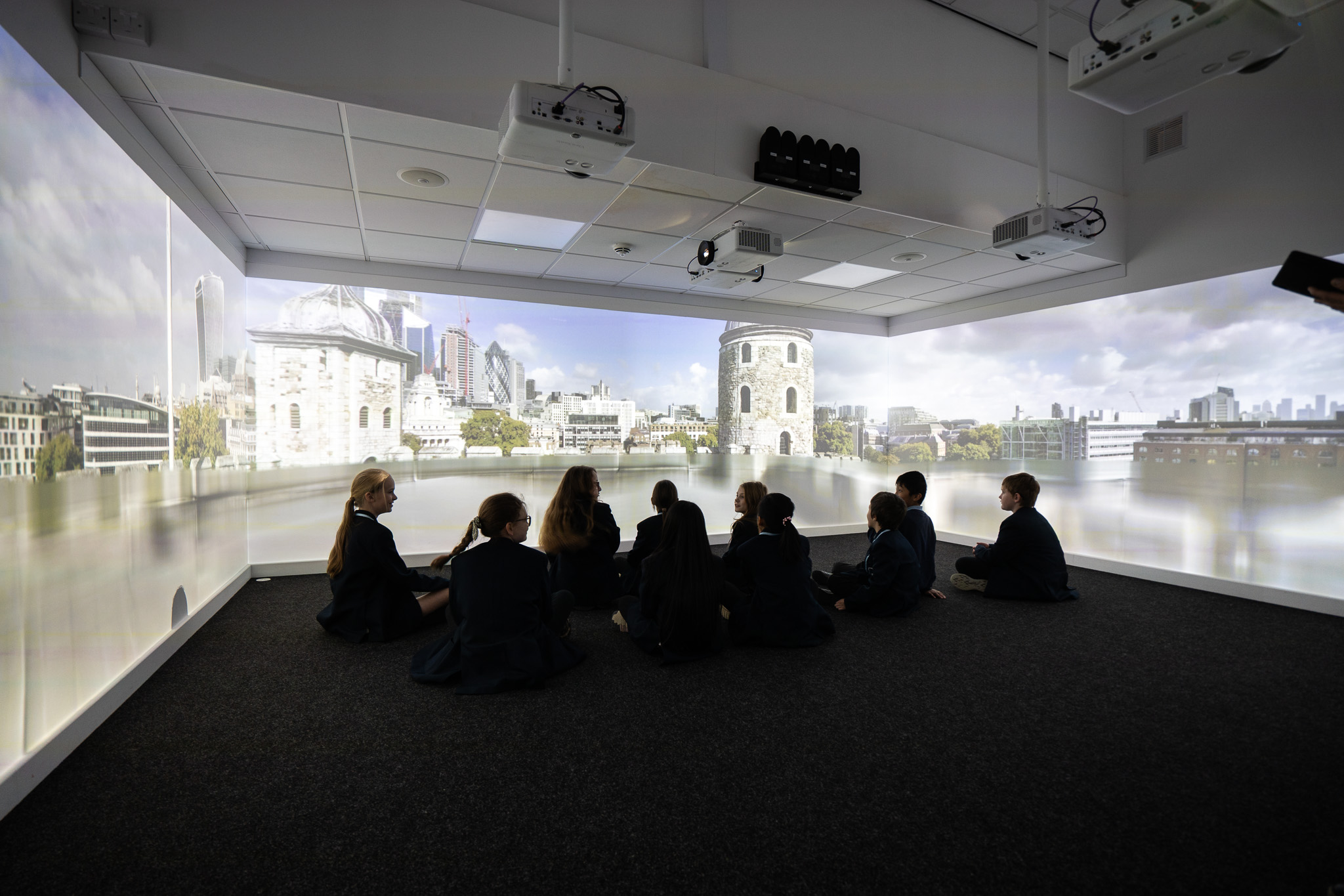 Students sit on a carpeted floor in a dark room, looking at a curved, immersive screen showing a city's skyline under a cloudy sky.