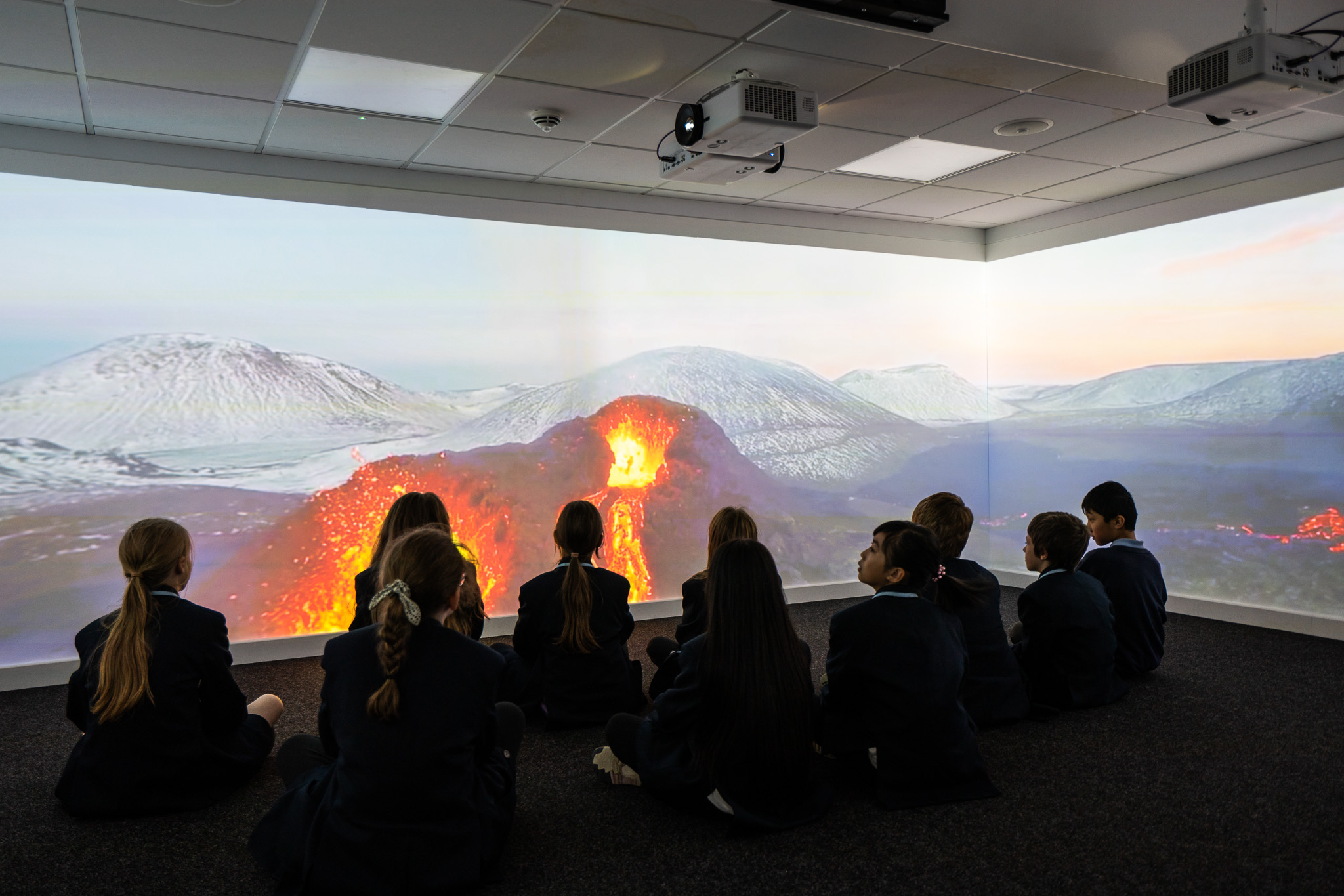 Children in school uniforms sit on the floor, captivated by a vibrant projection of an erupting volcano in a darkened room, creating an immersive learning atmosphere.