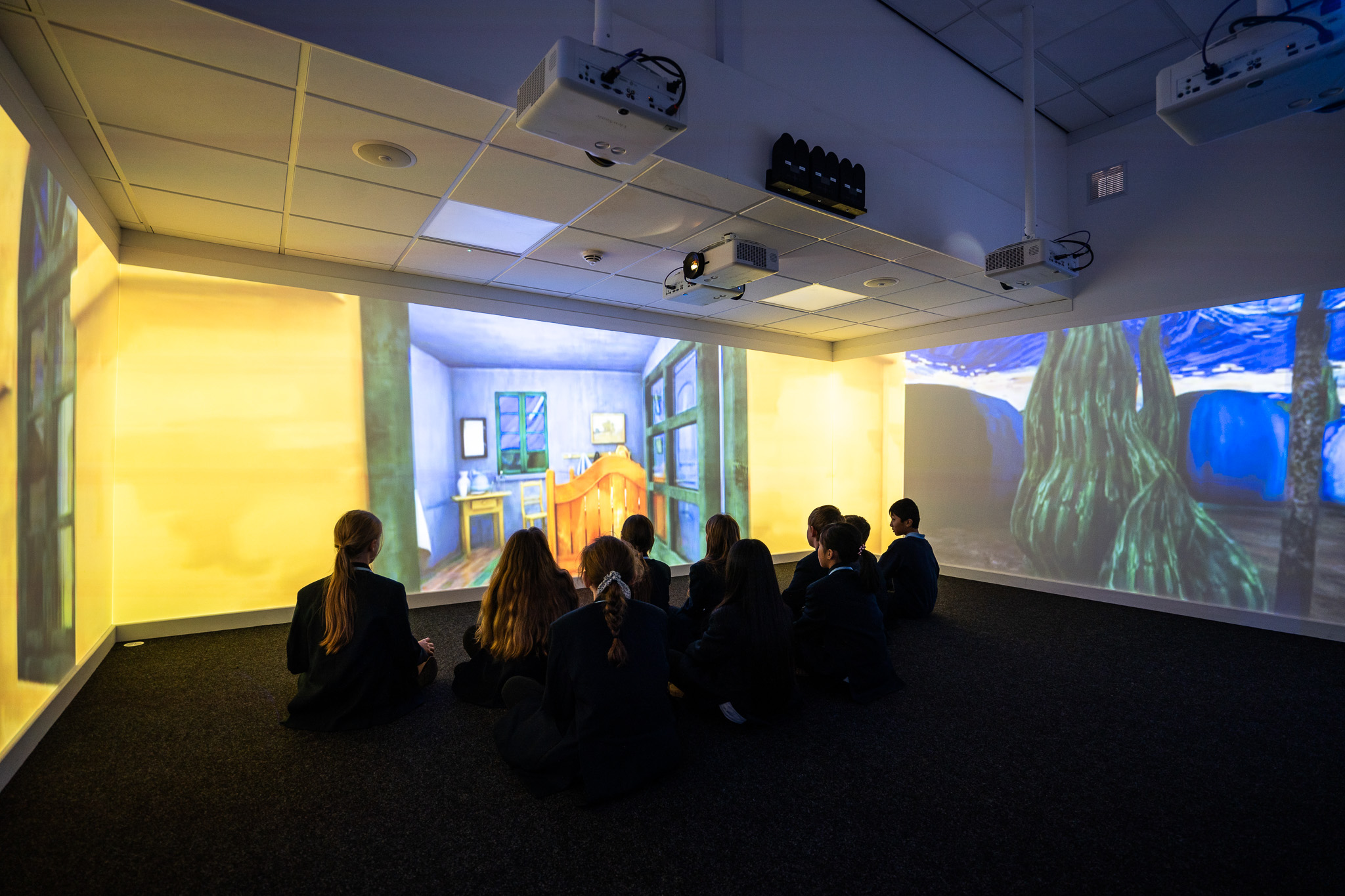 Students sit on the floor in a dim room, absorbed by vibrant digital projections of Van Gogh paintings on the walls, creating an immersive art experience.