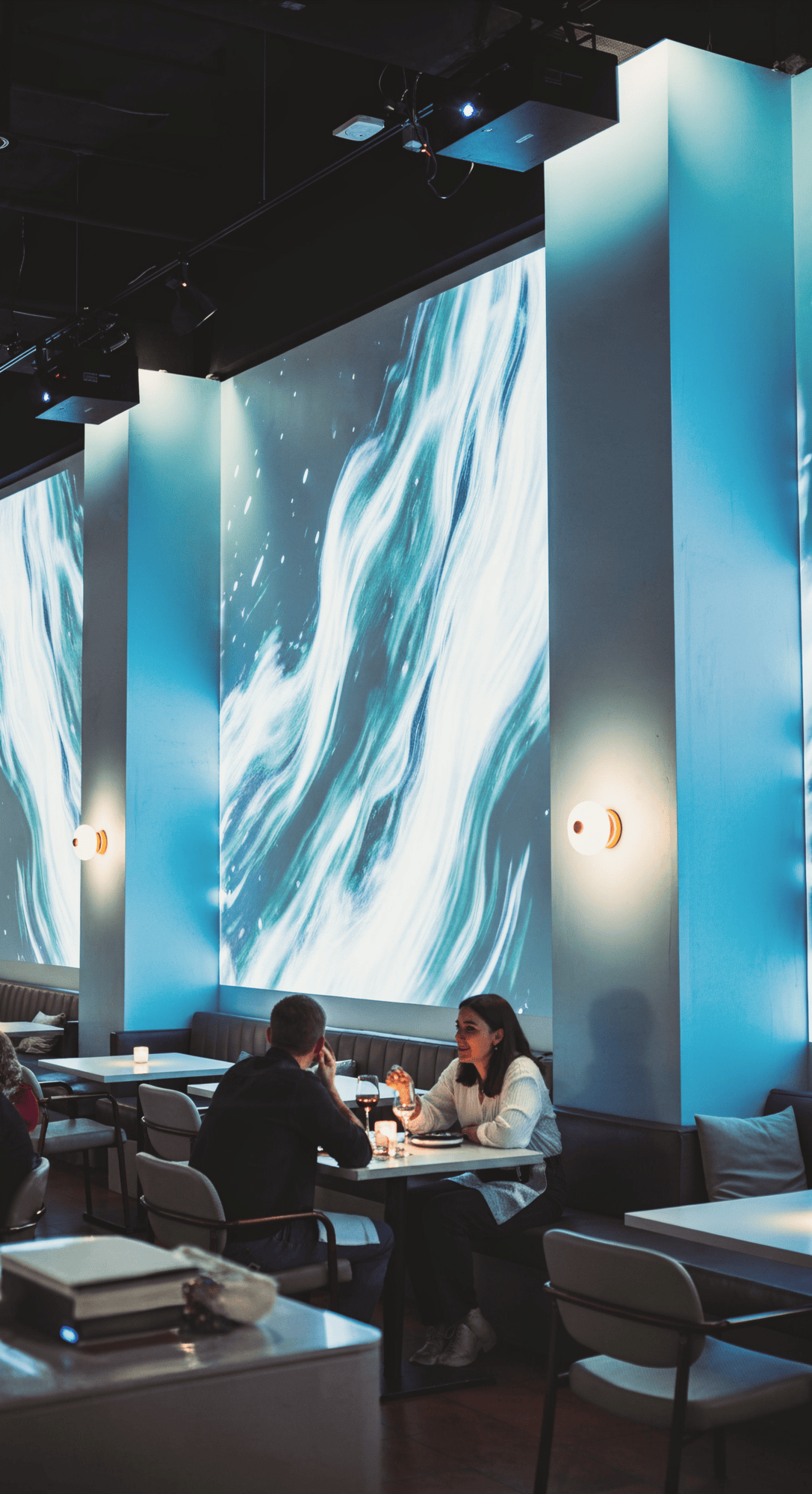 A cozy restaurant with two people seated at a table, engaged in conversation. Blue-lit walls display flowing wave patterns, creating a calming ambiance.