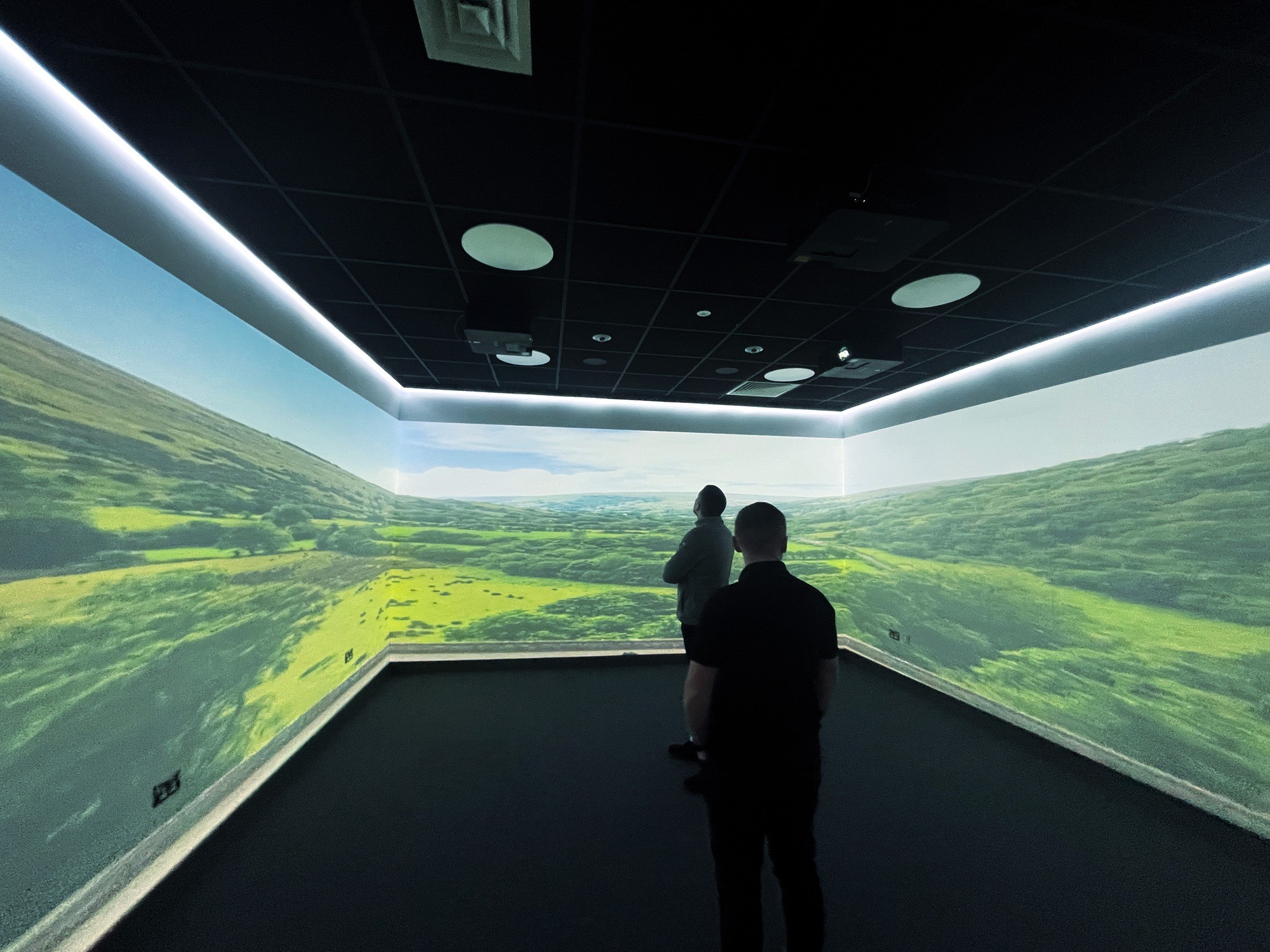 Two people stand in a dark room with large screens projecting a bright, lush landscape. The atmosphere is immersive and serene.