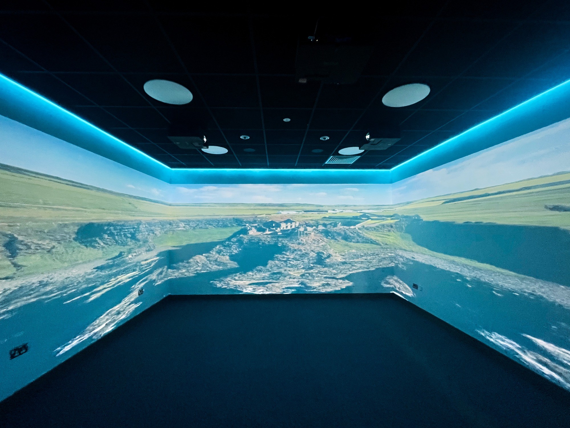 Room with 360-degree projection of a vibrant coastal landscape featuring cliffs and ocean. The ceiling has blue LED lights, creating an immersive experience.