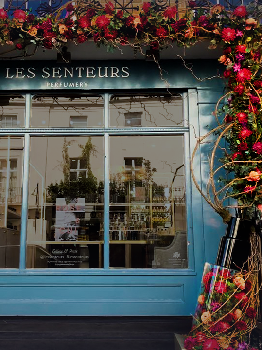Storefront of a perfumery with blue walls adorned with vibrant red and pink flower garlands. Large windows reflect nearby buildings, creating an inviting, elegant atmosphere.