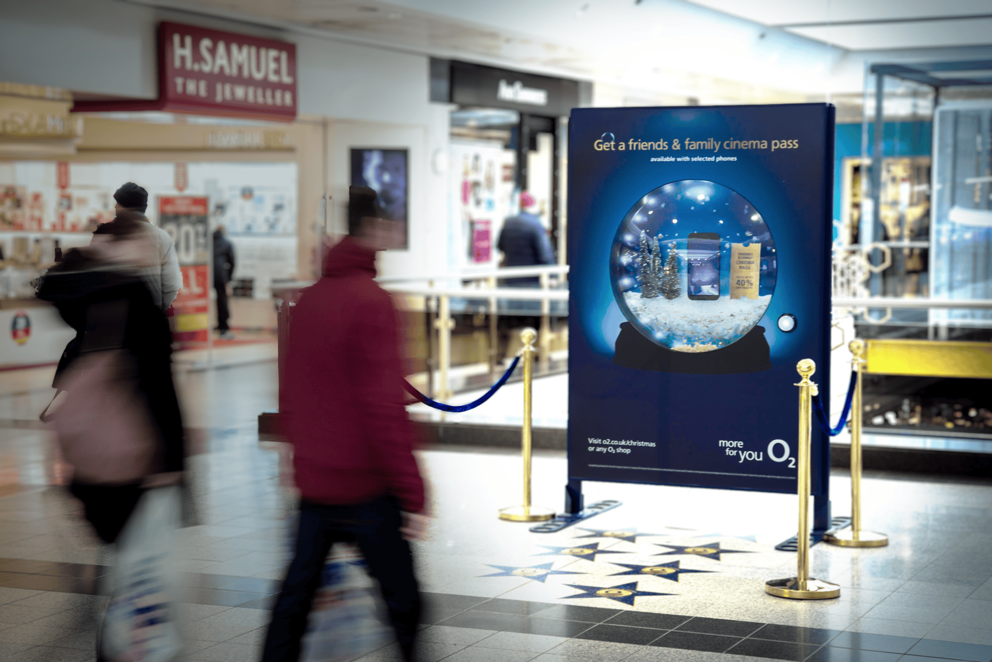 Busy shopping mall with blurred people walking past a large O2 advertisement. Sign offers a 