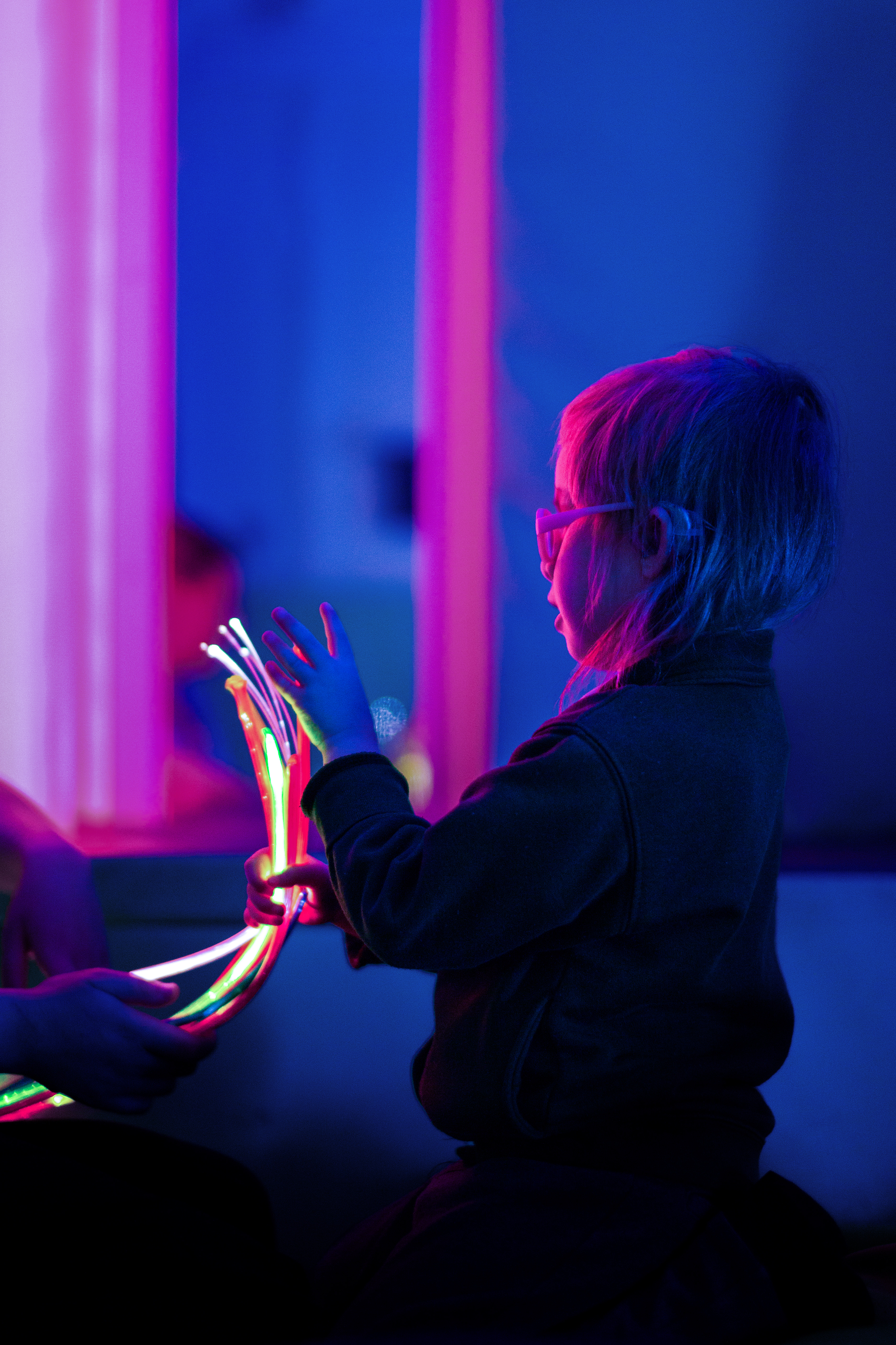 A child with glasses interacts with glowing fiber optic strands in a dimly lit room. The blue and pink ambient lighting creates a calming atmosphere.