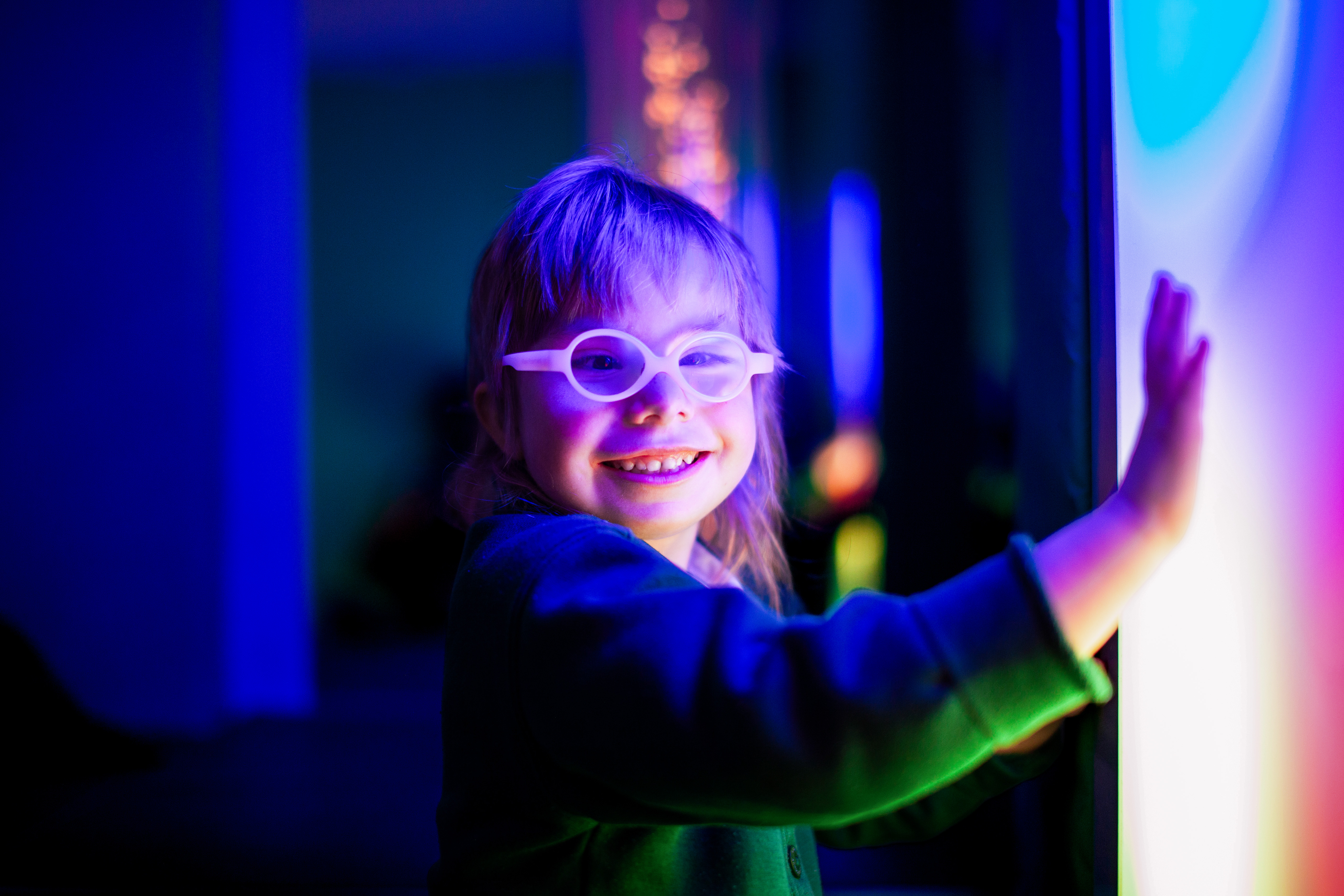 Young child wearing glasses smiles brightly, touching a glowing, colorful wall in a dark sensory room. The scene is vibrant and joyful.