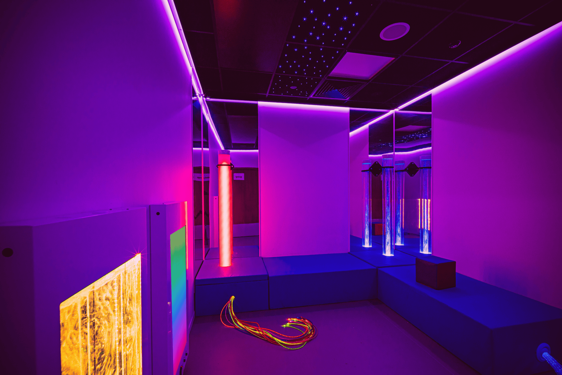 A sensory room with calming purple lighting, neon tubes, mirrored walls, and fiber optic cables on the floor. The atmosphere is soothing and futuristic.