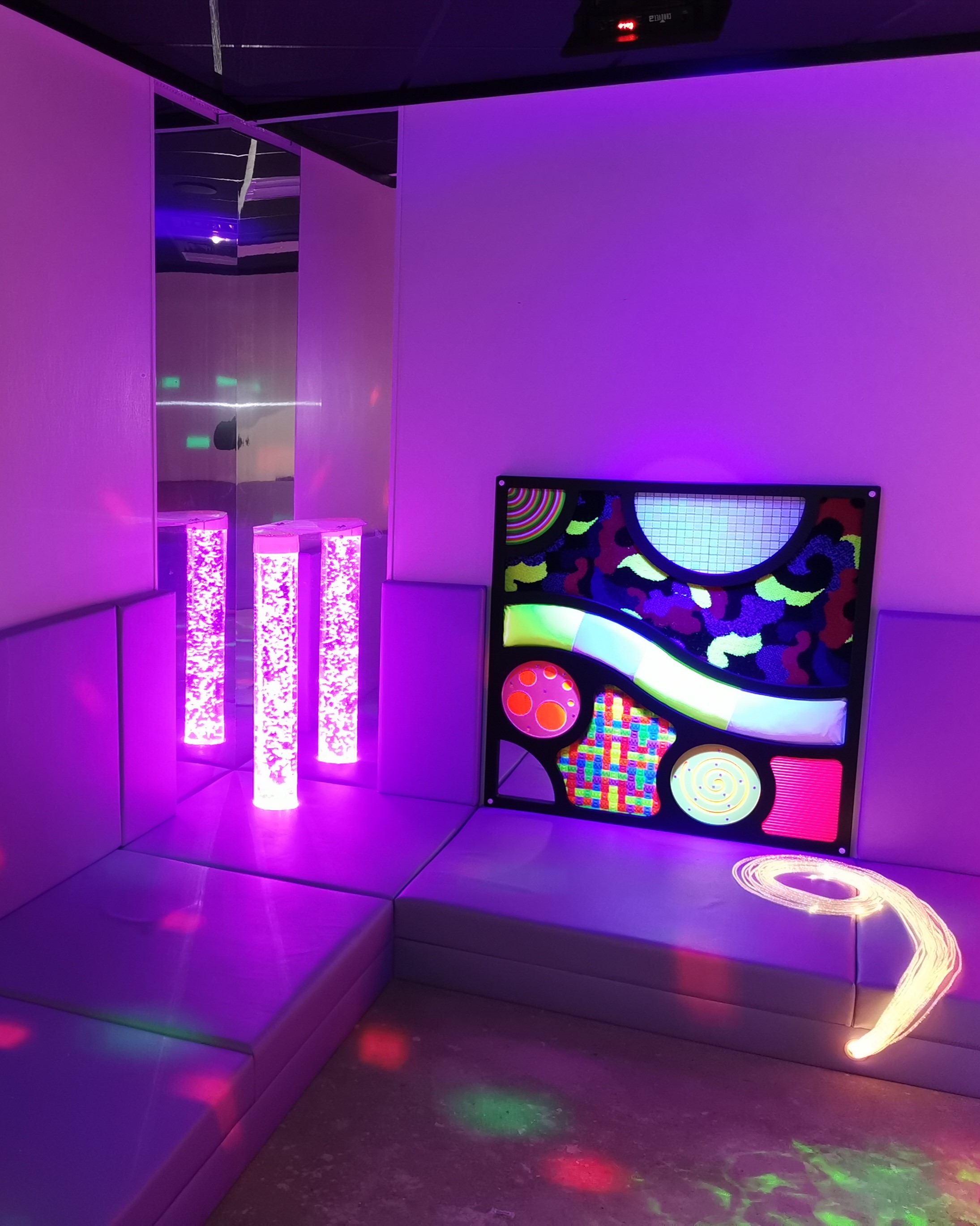 An aurora sensory room with glowing purple lights, cushioned walls, three bubble tubes, and a colorful abstract wall panel, creating a calming, immersive atmosphere.