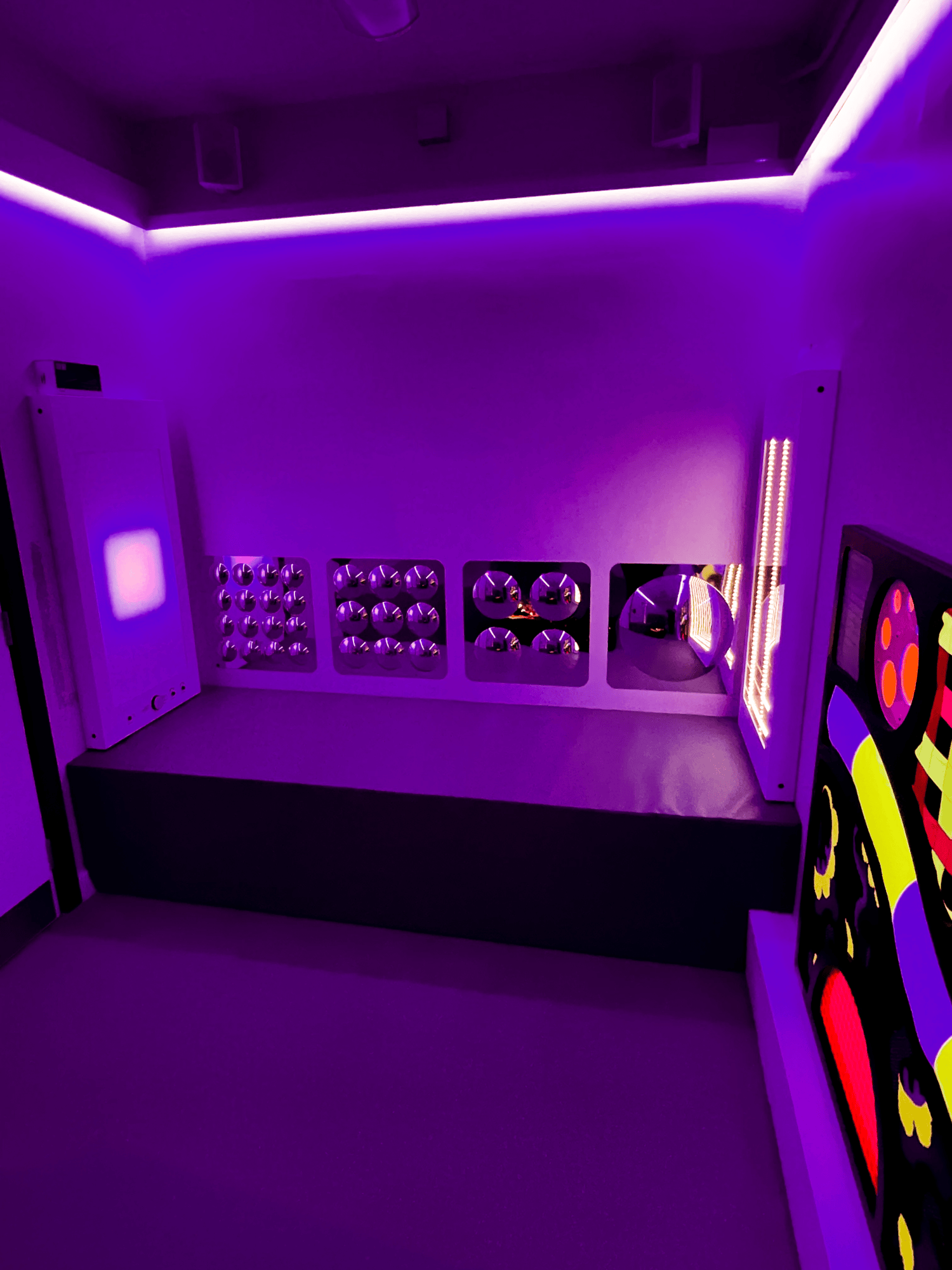 A sensory room bathed in purple light features tactile panels with various textures and patterns. It conveys a calming, interactive environment.