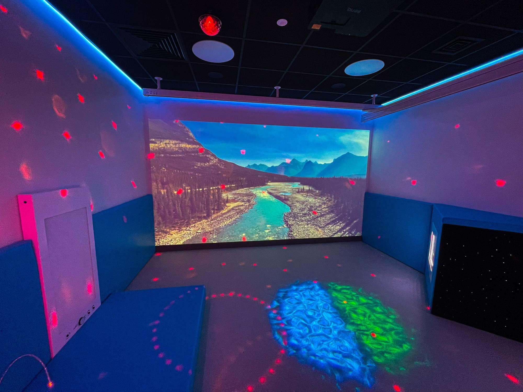 A sensory room features a large wall projection of a river and mountains. The space is dimly lit, with blue and red lights creating a calming ambiance.