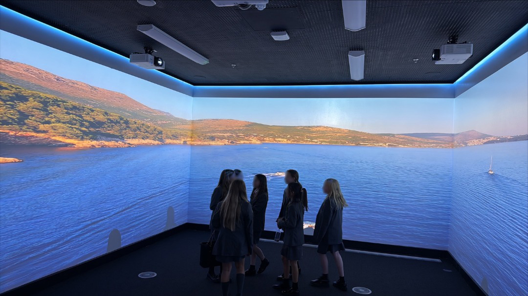 A group of students stands in a dark room with wraparound wall projections of a serene coastal landscape, featuring blue waters and scenic hills.