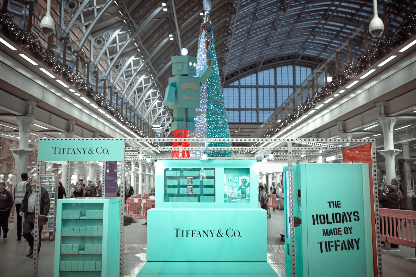 Tiffany & Co. holiday display in a station, featuring a large teal Tiffany box, a robot, and a sparkling tree under an arched glass roof. Festive and elegant.