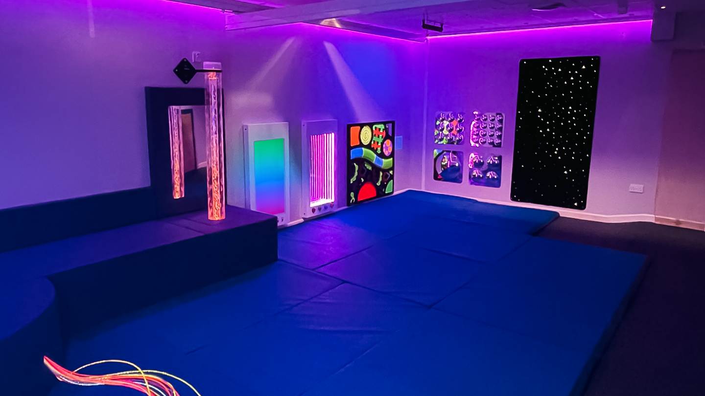 Neon-lit sensory room with blue padded flooring, glowing wall panels, fiber-optic light strands, and starry artwork creates a playful mood.