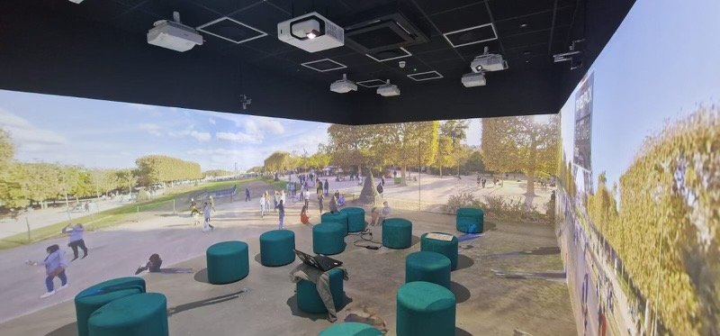 A room with multiple green ottomans and projection screens on the walls displays an outdoor park scene with people walking and trees in the background.