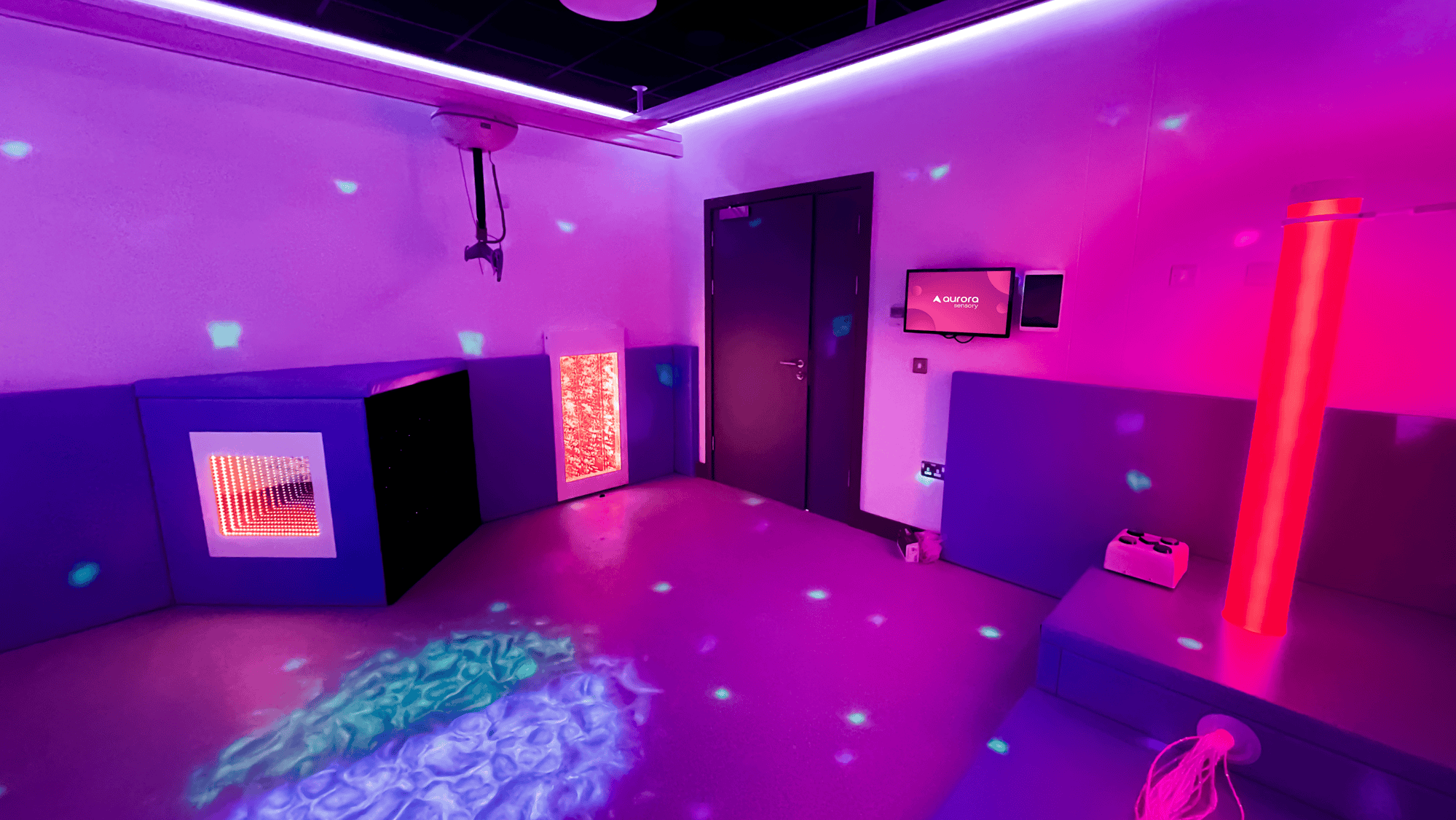 A sensory room with dim lighting and colourful projections on walls and floor. Features include a glowing bubble tube, interactive panels, and soft seating, creating a calming atmosphere.