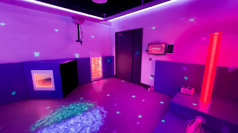 A sensory room with dim lighting and colourful projections on walls and floor. Features include a glowing bubble tube, interactive panels, and soft seating, creating a calming atmosphere.