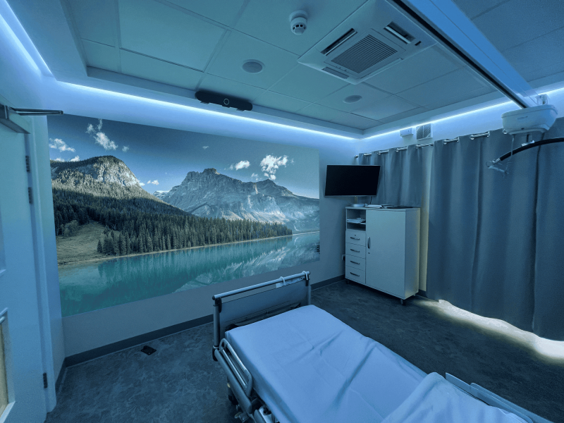 Hospital room with a serene atmosphere; features a bed, curtained window, and large wall projection of a tranquil mountain lake under a clear blue sky.