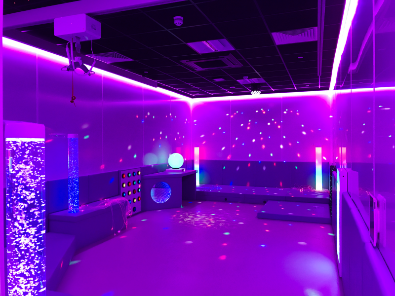 sensory room glowing with purple and blue neon, starry projections, light columns, glowing orbs, and padded seating; playful, calming mood.