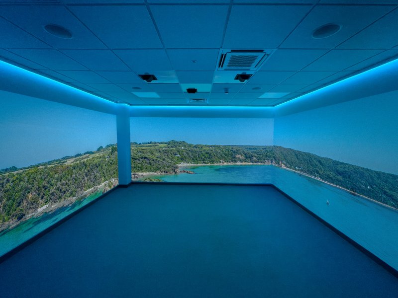 Blue-lit immersive room with panoramic projection showing a lush green island, rocky coastline, and calm turquoise bay beneath a clear sky.