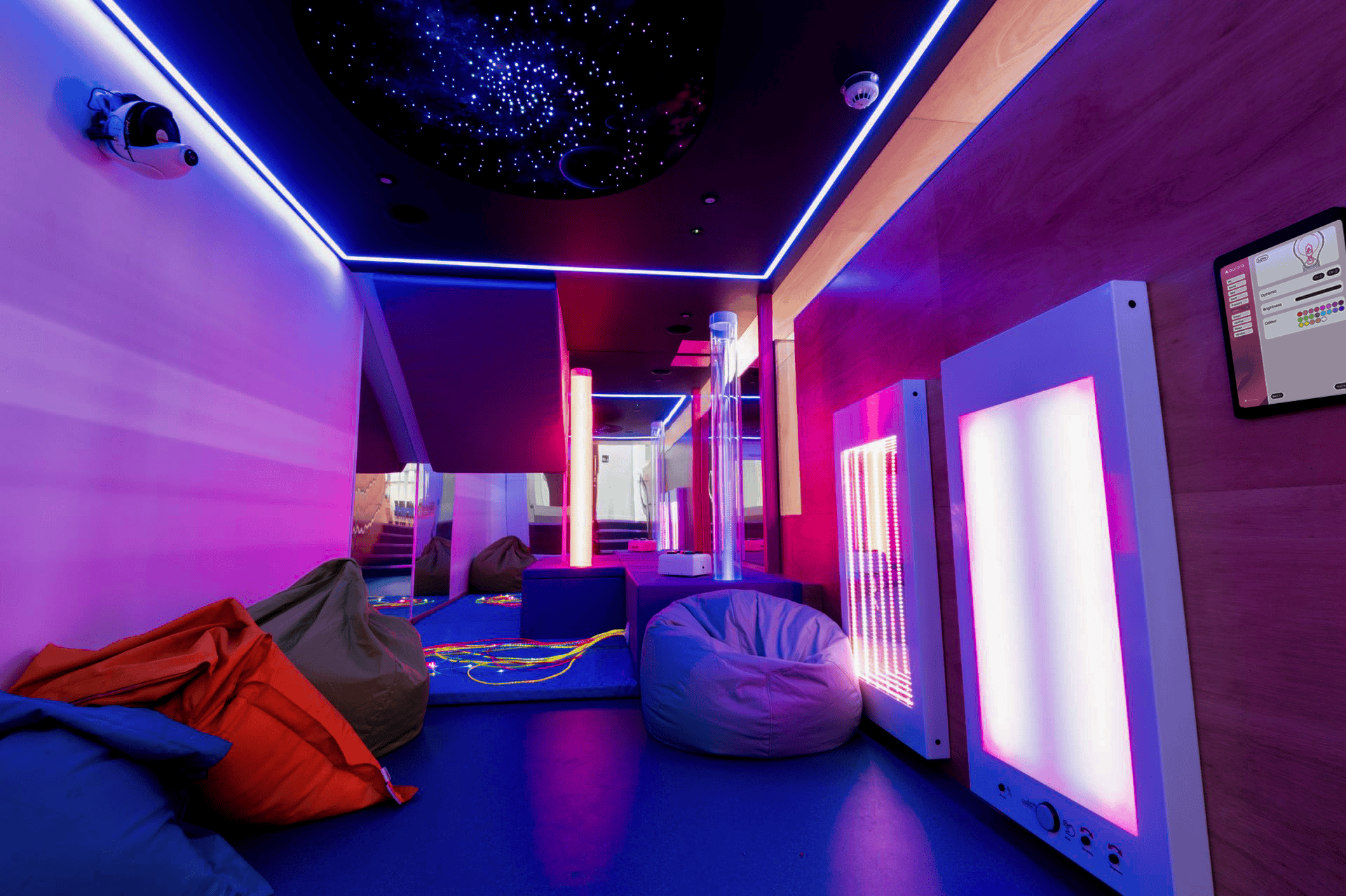 A sensory room with dim neon lighting in purple and blue hues. Bean bags and calming LED panels create a relaxing atmosphere. Ceiling resembles a starry sky.