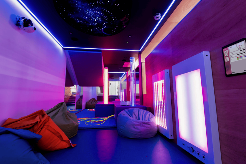 A sensory room with dim neon lighting in purple and blue hues. Bean bags and calming LED panels create a relaxing atmosphere. Ceiling resembles a starry sky.
