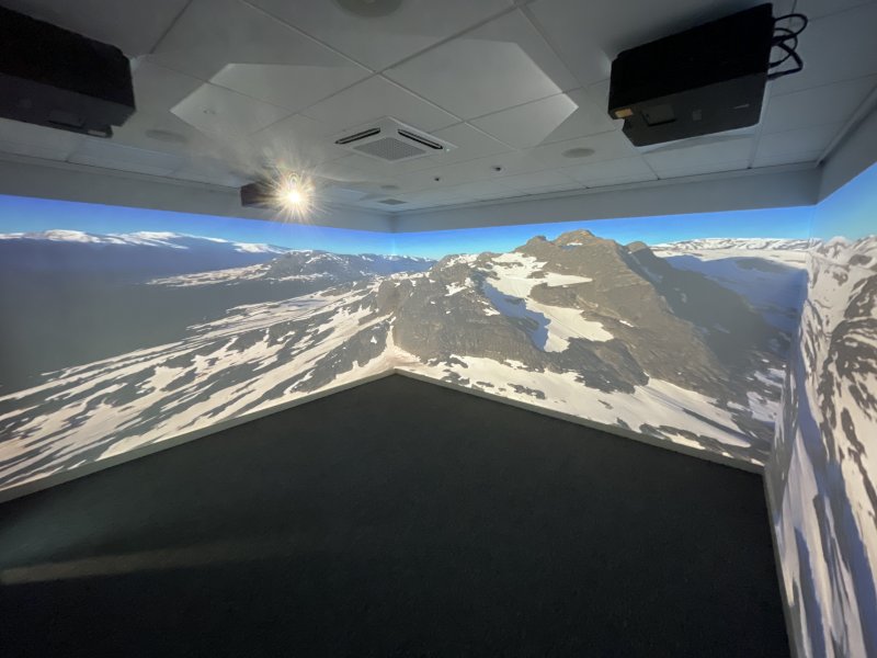 Immersive room projection shows a sweeping, snow-covered mountain range beneath blue skies, creating a dramatic, awe-inspiring atmosphere.