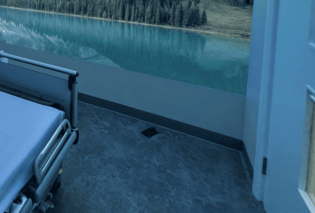 Hospital room with a white bed, grey curtains, hidden ambient coloured lighting and a single wall aurora projection image of a nature mural of mountains and a lake creates a calming atmosphere.
