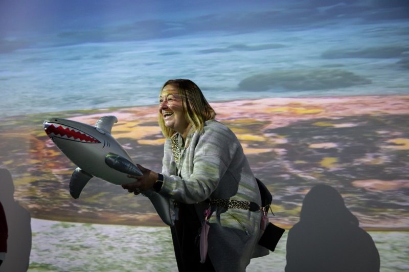 Smiling woman holds a model shark before a colorful ocean projection, creating a playful, immersive educational scene.