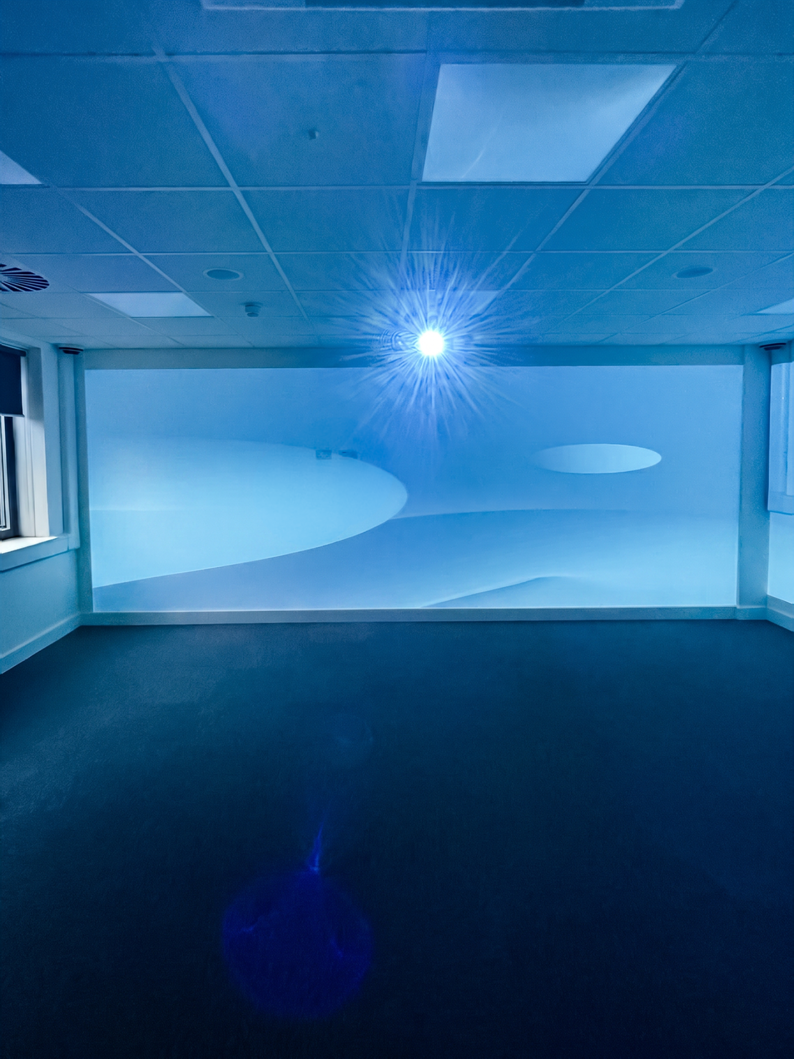 Blue-lit empty room with an abstract wave-pattern projected onto a wall, bright ceiling light flare, and dark carpet, creating a cool, futuristic mood.