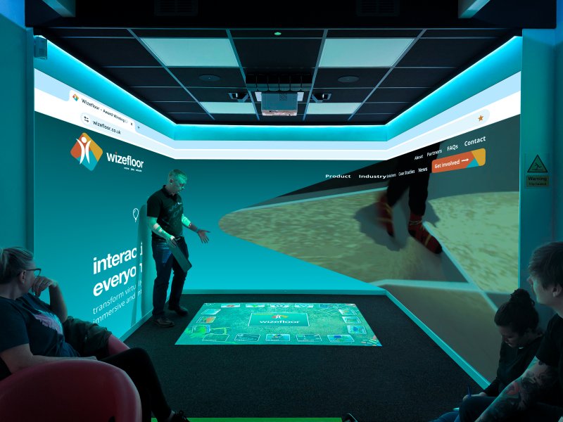 People explore an immersive Wizefloor room surrounded by turquoise maps and screens; one participant stands on an interactive floor surrounded by projected visuals