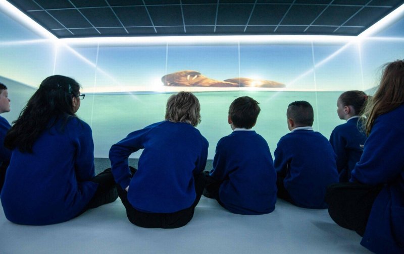 Schoolchildren in blue uniforms sit together surrounded by immersive projection of an underwater scene, creating wonder.