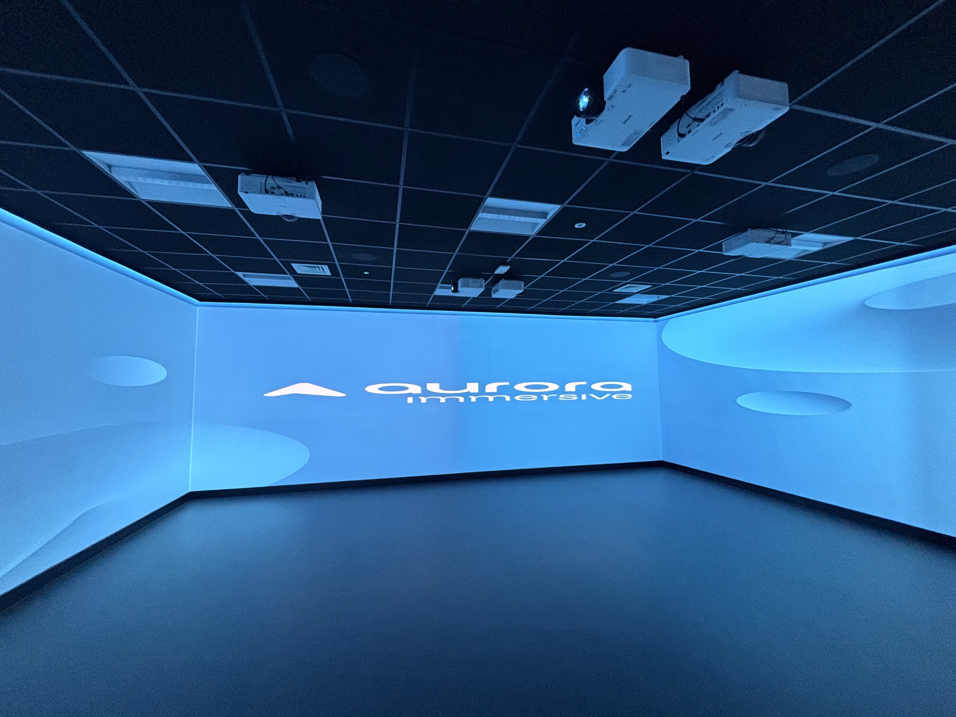 Empty immersive projection room bathed in blue light, with ceiling-mounted projectors, curved walls, and a glowing abstract logo; futuristic, calm mood.