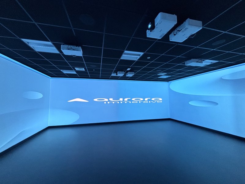 Empty immersive projection room bathed in blue light, with ceiling-mounted projectors, curved walls, and a glowing abstract logo; futuristic, calm mood.