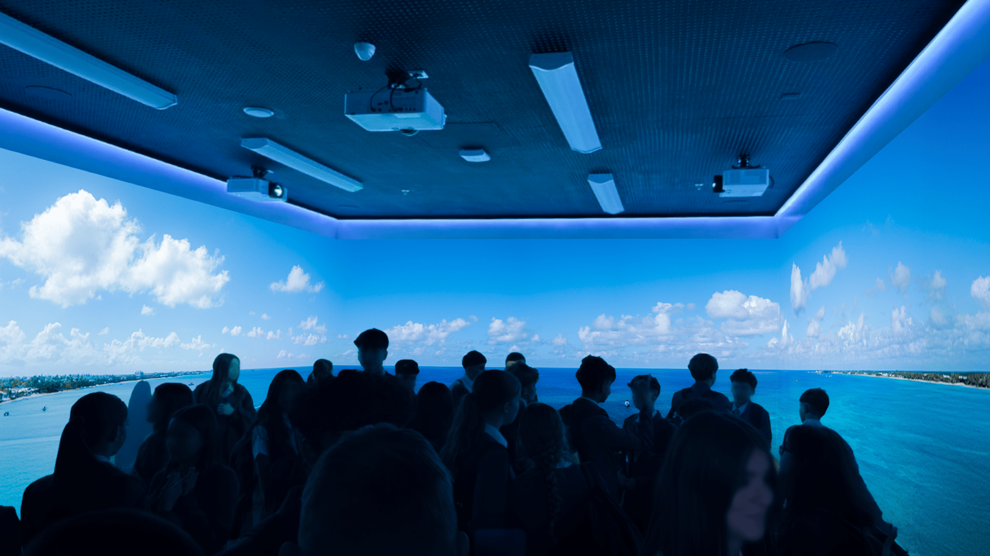 A group of people in silhouette stands inside a room with panoramic projections of an ocean view and blue sky, creating a serene atmosphere.