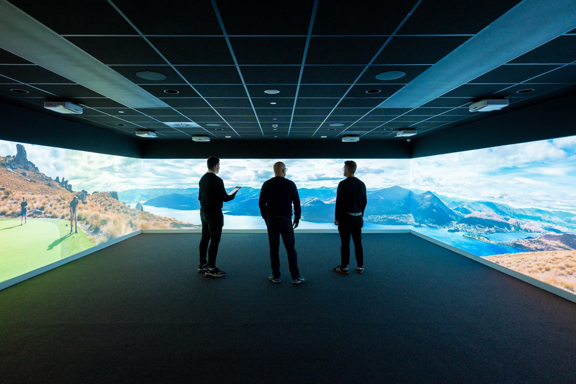 Three people stand in a dark room with panoramic landscape projections of mountains and a lake, examining the immersive digital display.